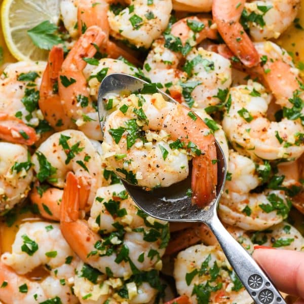 Cooked shrimp scampi without wine with parsley, lemon, and garlic, on a round platter, with a spoon lifting out a single shrimp.