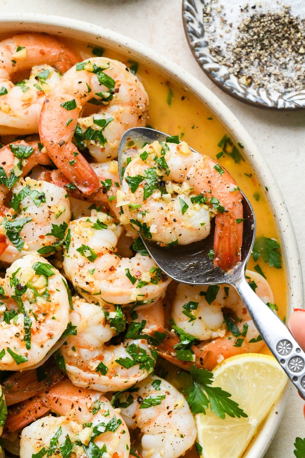 Close up of cooked shrimp scampi without wine with parsley, lemon, and garlic, on a round platter, with a spoon lifting out a single shrimp from the edge of the plate.