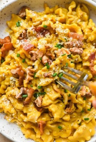 Creamy, dairy free butternut squash pasta with bacon, walnuts, and chives, in a shallow speckled ceramic bowl with a fork piercing a piece of pasta to show the creamy texture of the pasta sauce.