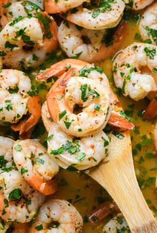 A wooden spatula lifting out several shrimp from prepared skillet of shrimp scampi.