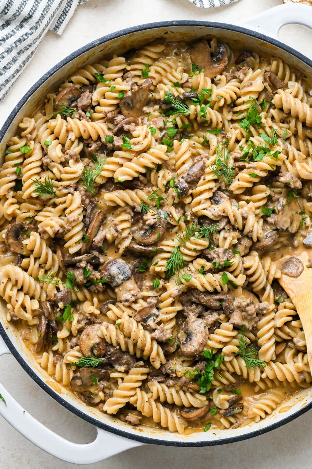 Dairy free beef stroganoff with gluten free fusili noodles and mushrooms in a large, white, enameled cast iron skillet, garnished with fresh dill and fresh parsley, on a creamy off white background.