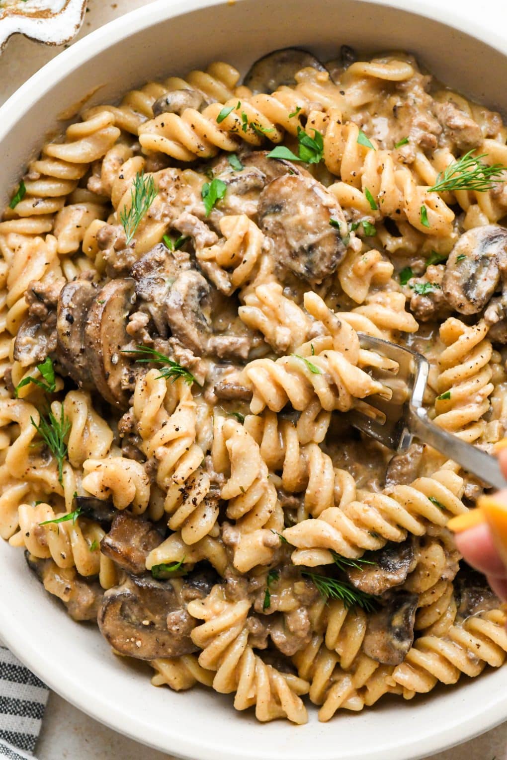 A shallow ceramic bowl of dairy free beef stroganoff with ground beef and mushrooms, garnished with fresh dill and fresh parsley, with a fork digging into the pasta for a creamy bite.