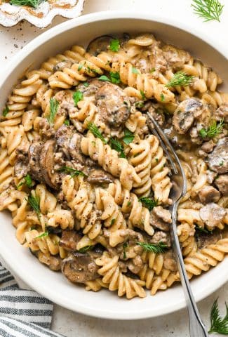 A shallow ceramic bowl of dairy free beef stroganoff with ground beef and mushrooms, garnished with fresh dill and fresh parsley, and a fork angled into the bowl to reveal some of the creamy texture of the sauce.