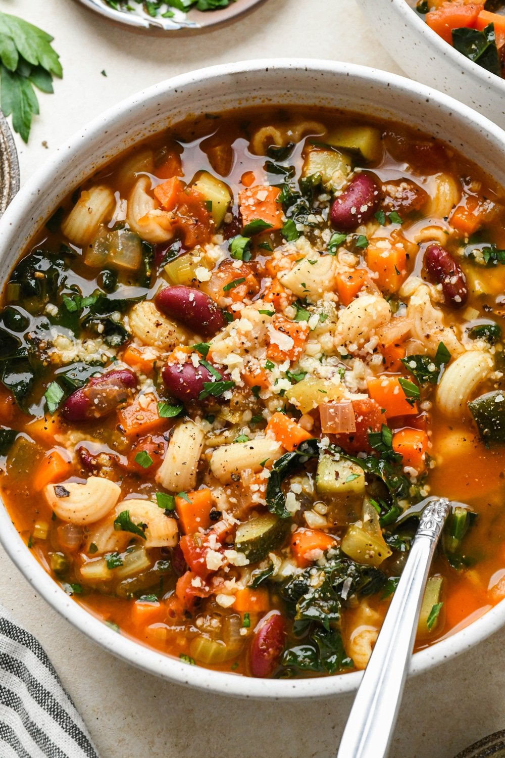 Gluten free minestrone soup in a large soup bowl topped with fresh parsley and grated parmesan, with a spoon angled into the bowl.
