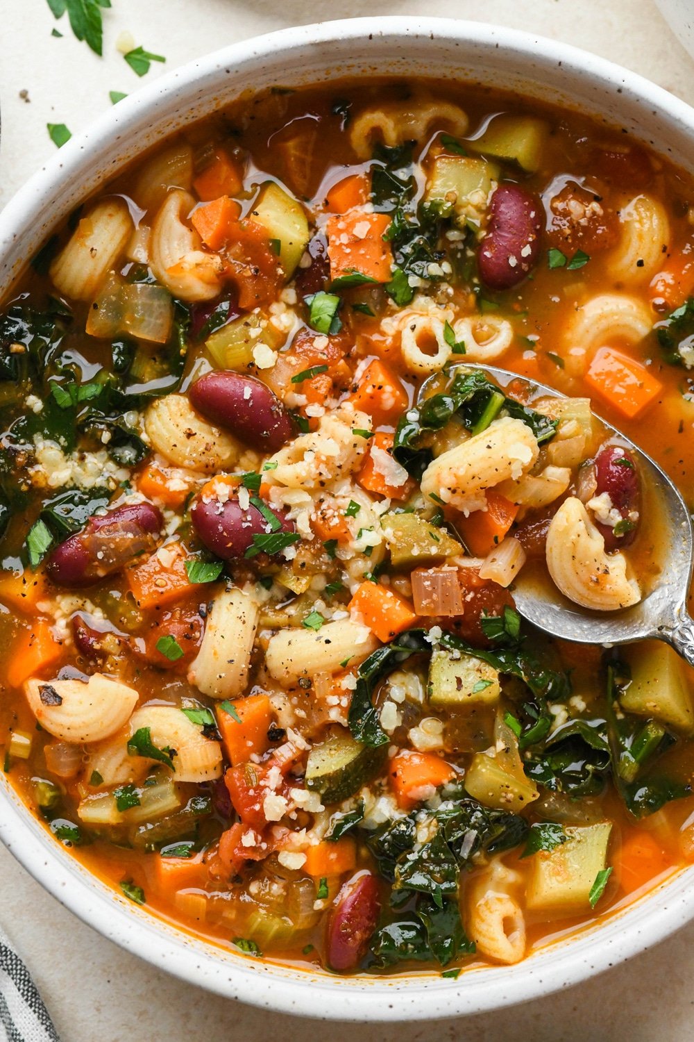 Gluten free minestrone soup in a large soup bowl topped with fresh parsley and grated parmesan, with a spoon lifting out a bite of the soup to show the ingredients.