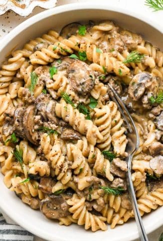 A shallow ceramic bowl of dairy free beef stroganoff with ground beef and mushrooms, garnished with fresh dill and fresh parsley, and a fork angled into the bowl to reveal some of the creamy texture of the sauce.