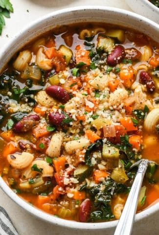 Gluten free minestrone soup in a large soup bowl topped with fresh parsley and grated parmesan, with a spoon angled into the bowl.