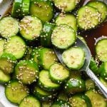 Prepared cucumber sesame salad in a shallow white speckled ceramic serving bowl, topped with sesame seeds, with a spoon lifting out several pieces of cucumber.