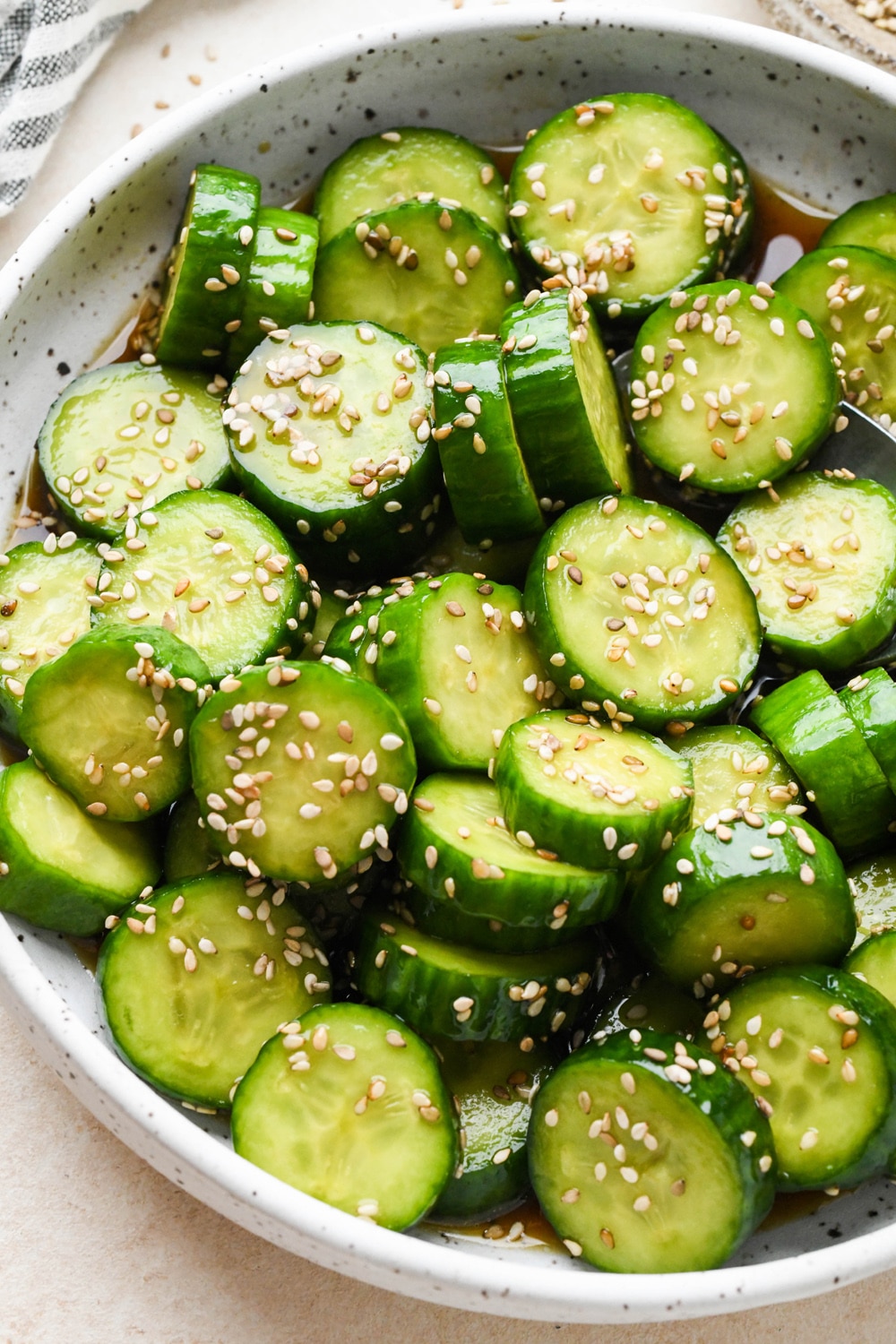 Angled view of prepared cucumber sesame salad in a shallow white speckled ceramic serving bowl, topped with sesame seeds.
