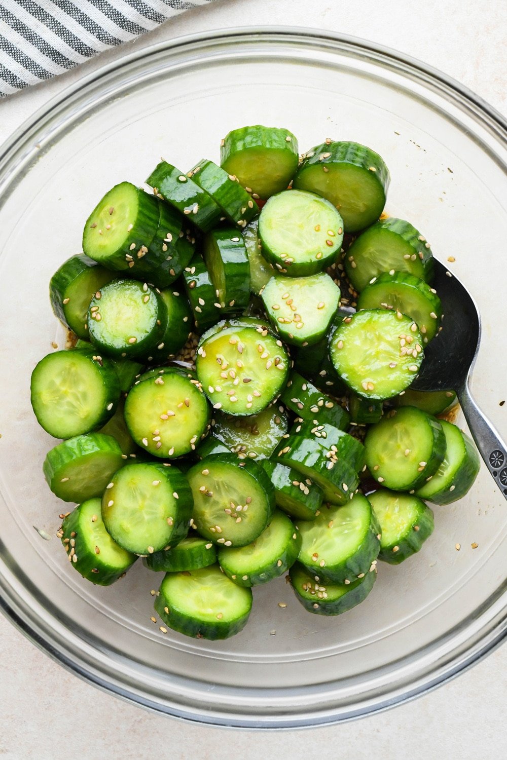 How to make cucumber sesame salad: Dressing poured over sliced cucumbers in a medium sized glass bowl, before marinating.