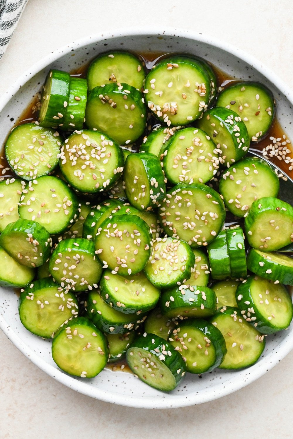 How to make cucumber sesame salad: Dressing poured over sliced cucumbers in a shallow ceramic serving bowl, after marinating and topping with additional sesame seeds.