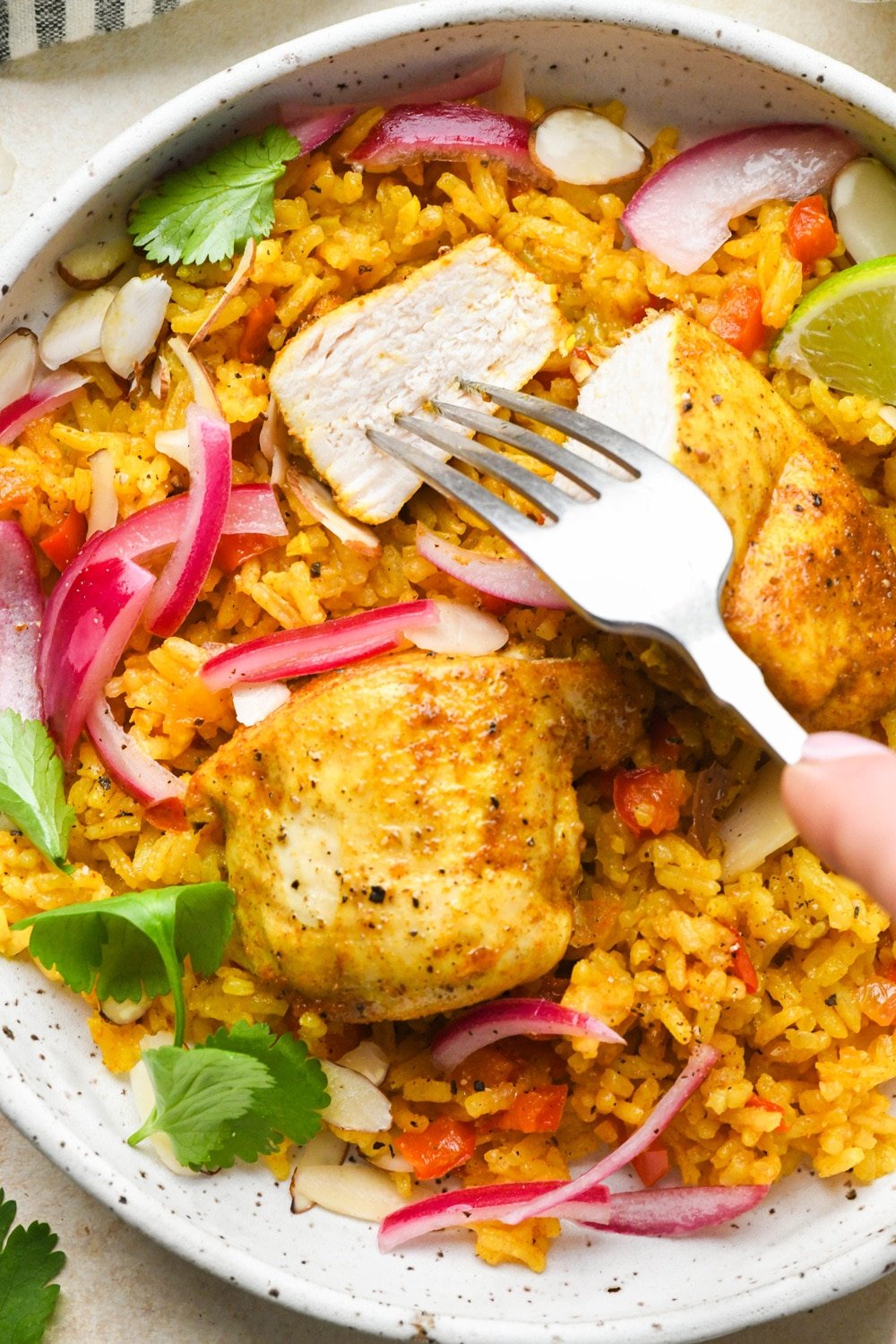 A single serving of vibrantly colored baked curry chicken and rice in a shallow speckled bowl topped with pink pickled red onions, cilantro, sliced almonds, and a lime wedge. A fork is piercing a cut bite of the chicken breast.