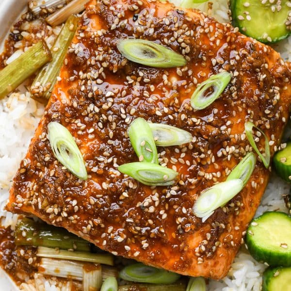 Baked garlic ginger salmon garnished with freshly sliced green onion, on top of white rice, next to a serving of sesame cucumber salad.