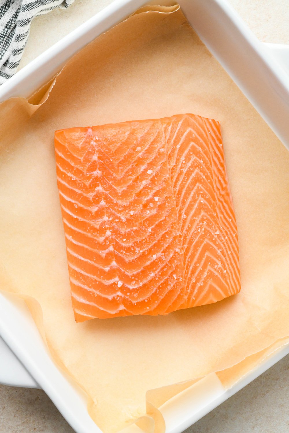 How to make garlic ginger salmon: Salted salmon filet in a small white square baking dish lined with parchment paper.