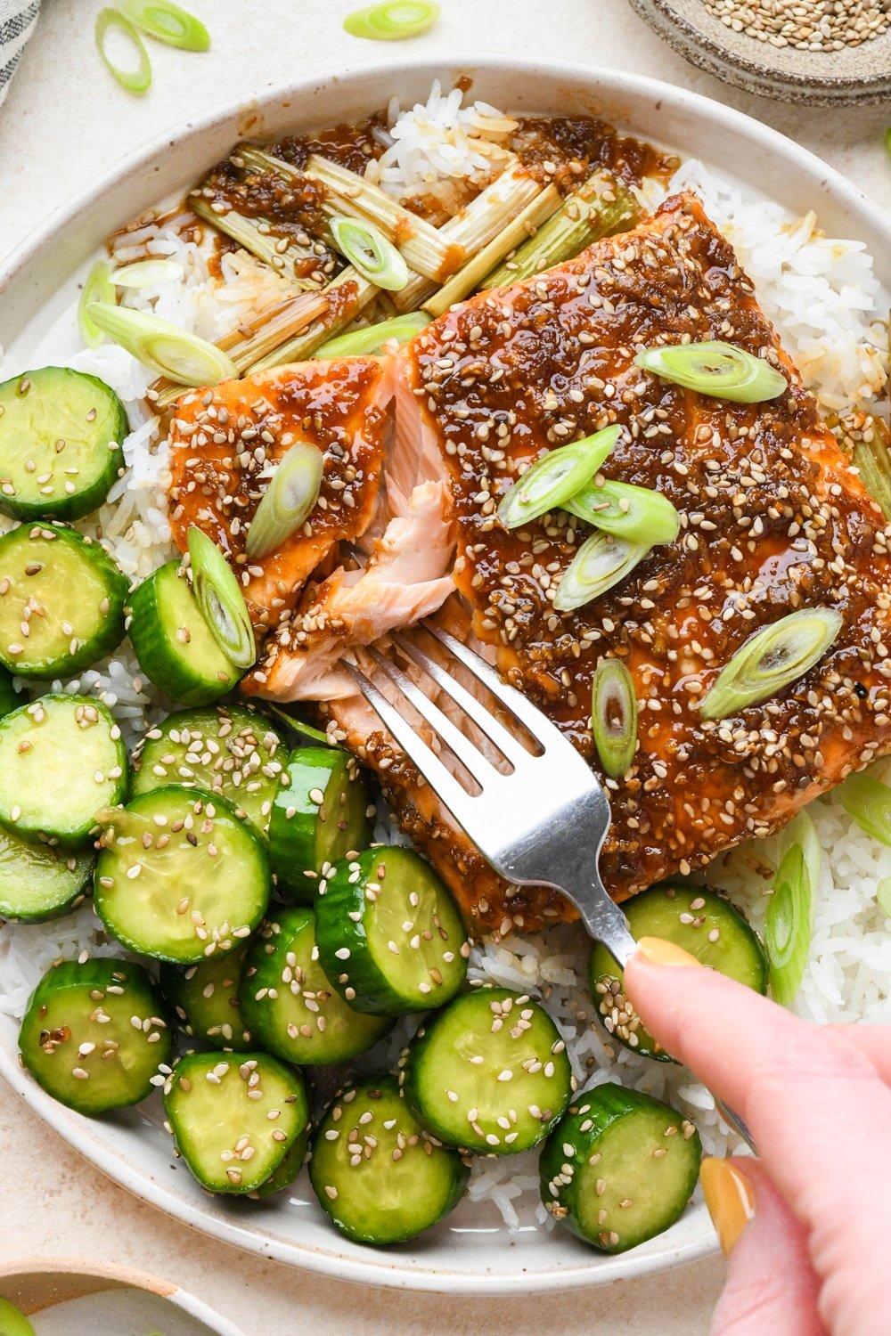 Caramelized garlic ginger salmon filet on white rice with a side of crunchy sesame cucumber salad, on a wide ceramic plate, with a fork digging into the salmon to show the tender interior.