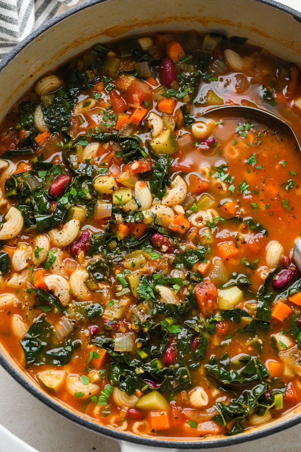 How to make Gluten Free Minestrone Soup: Cooked macaroni pasta added to soup before stirring to combine, with a ladle dipping into the soup pot to show the ingredients.