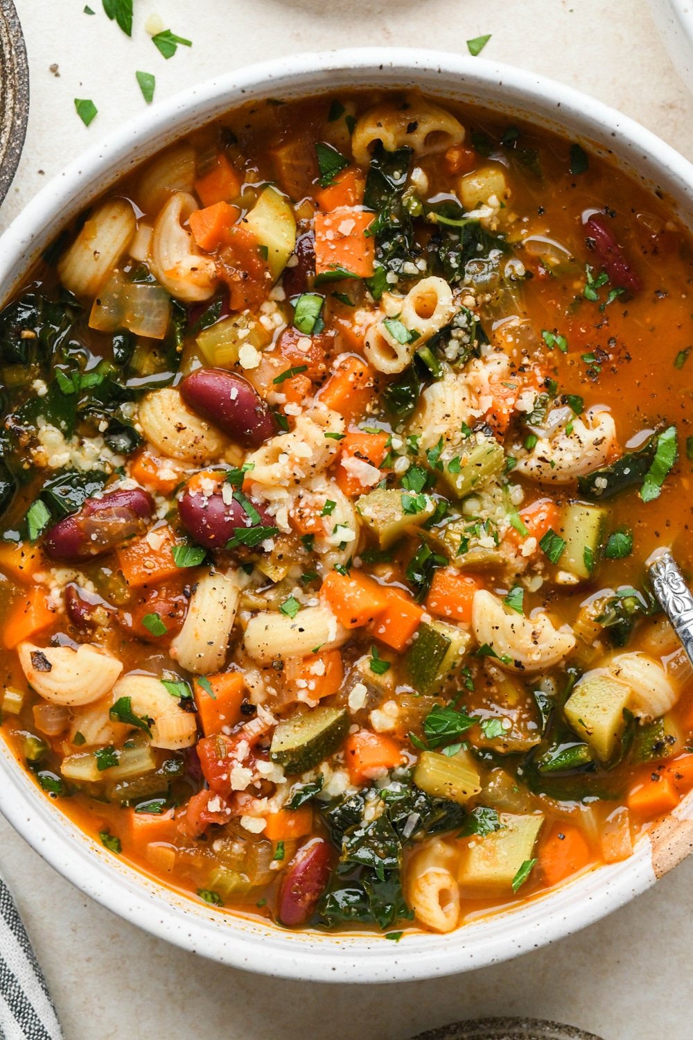 Gluten free minestrone soup in a large soup bowl topped with fresh parsley and grated parmesan, with a spoon lifting out a bite of the soup to show the ingredients.