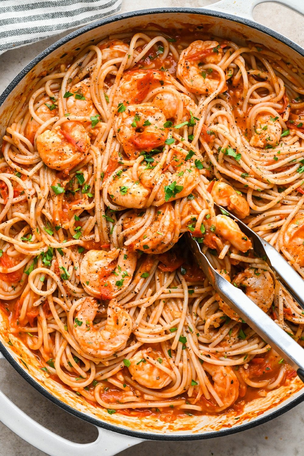 Burst cherry tomato and shrimp pasta fully prepared in a large enameled skillet, with kitchen tongs reaching into the pasta to pull out a serving.