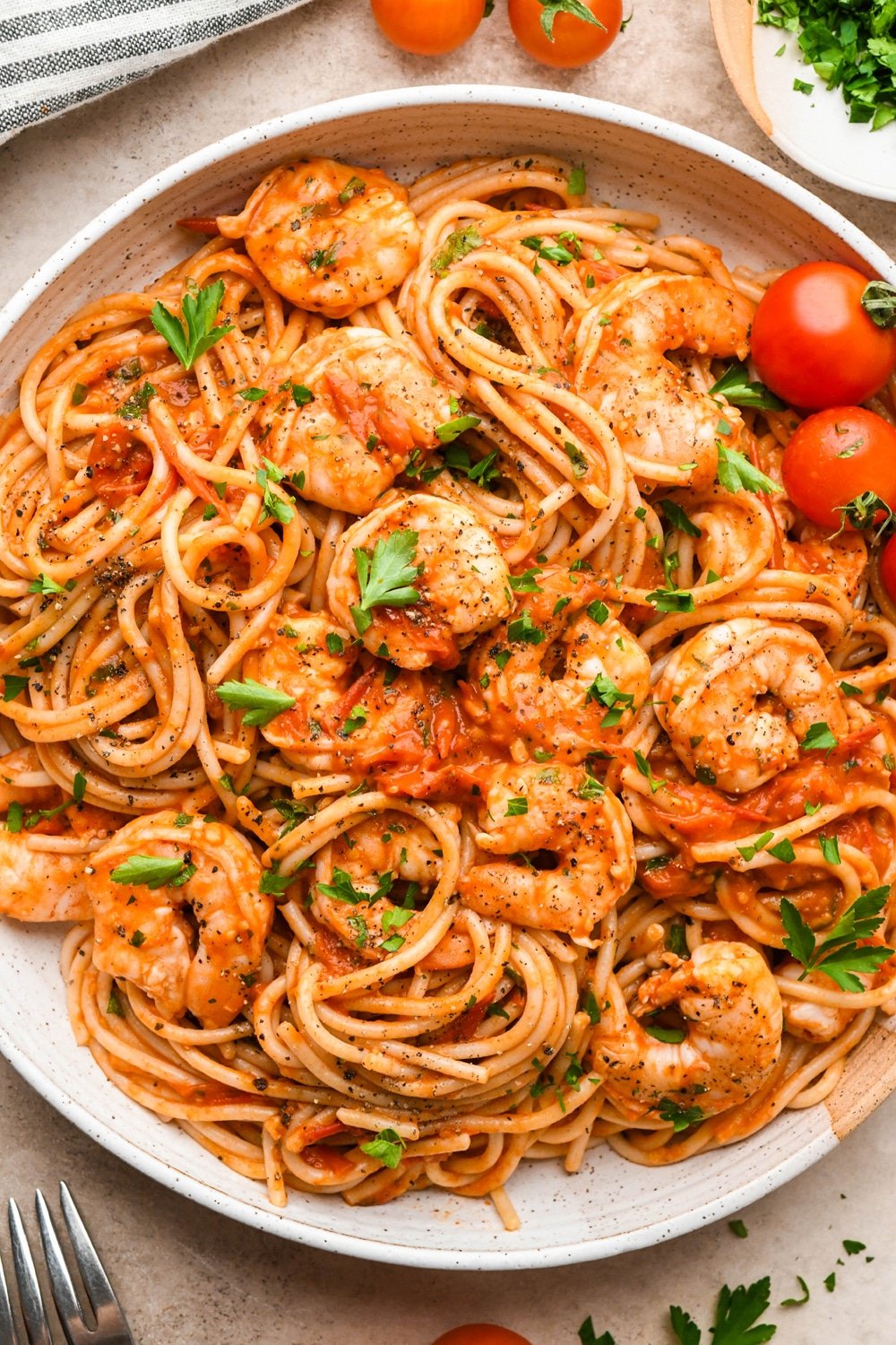 A large shallow ceramic bowl with a generous portion of burst cherry tomato and shrimp pasta. It is garnished with fresh parsley and there are several cherry tomatoes on the vine surrounding the bowl.