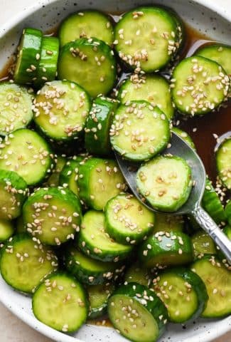 Prepared cucumber sesame salad in a shallow white speckled ceramic serving bowl, topped with sesame seeds, with a spoon lifting out several pieces of cucumber.