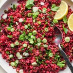 Quinoa and beet salad in a large shallow ceramic serving dish. The salad is garnished with sliced green onion, fresh chopped parsley, and lemon wedges on the side. A spoon is dipping into the salad to show the texture.