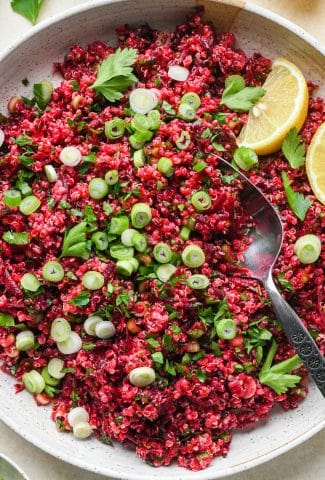 Quinoa and beet salad in a large shallow ceramic serving dish. The salad is garnished with sliced green onion, fresh chopped parsley, and lemon wedges on the side. A spoon is dipping into the salad to show the texture.
