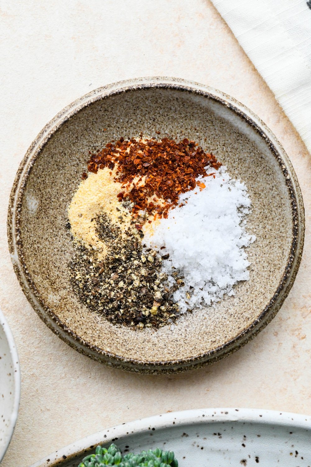 Garlic powder, chili flakes, kosher salt, and freshly ground black pepper in a small shallow ceramic pinch dish on a cream colored background.