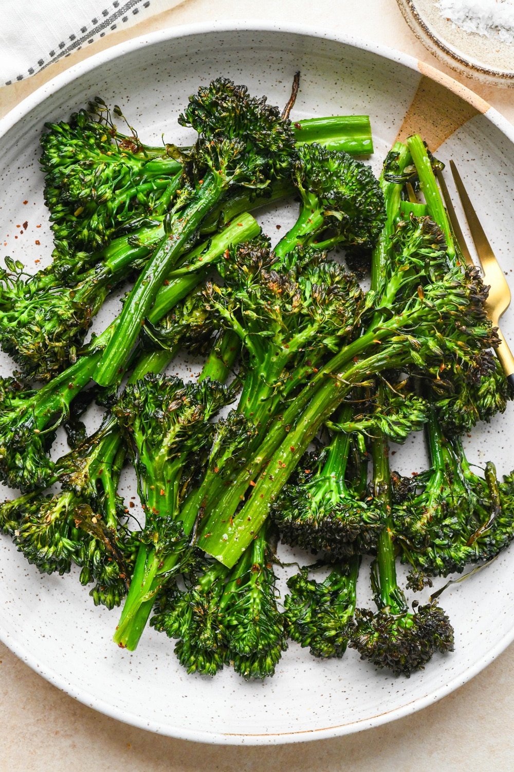 Roasted broccolini stalks in a shallow speckled ceramic serving bowl. The broccolini is bright green with crispy bits on the florets.