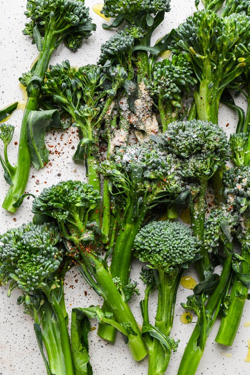 How to make roasted broccolini: Prepped stalks of broccolini on a sheet pan topped with olive oil and spices, before tossing to coat.