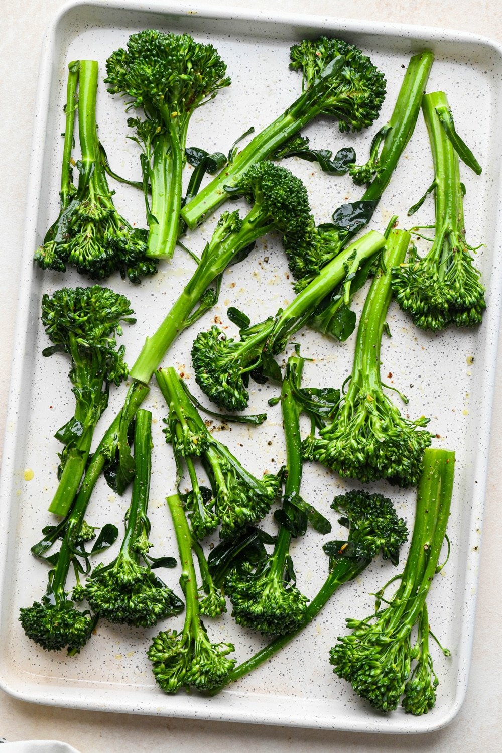 How to make roasted broccolini: Prepped stalks of broccolini on a sheet pan topped with olive oil and spices, before tossing to coat.