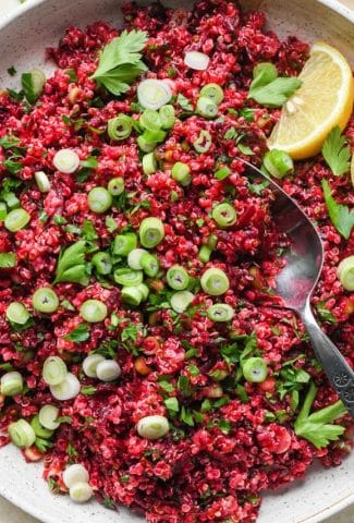 Quinoa and beet salad in a large shallow ceramic serving dish. The salad is garnished with sliced green onion, fresh chopped parsley, and lemon wedges on the side. A spoon is dipping into the salad to show the texture.