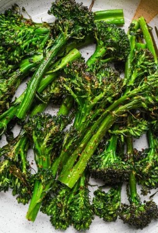 Roasted broccolini stalks in a shallow speckled ceramic serving bowl. The broccolini is bright green with crispy bits on the florets.