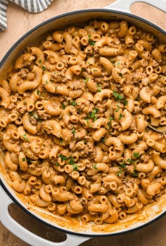 An off white ceramic coated cast iron skillet filled with dairy free and gluten free hamburger helper with ground beef and a creamy sauce topped with fresh parsley.