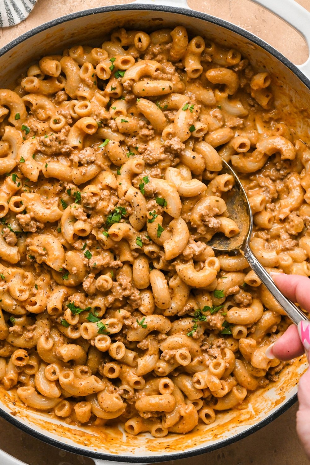 An off white ceramic coated cast iron skillet filled with dairy free and gluten free hamburger helper with ground beef and a creamy sauce topped with fresh parsley, and a spoon lifting some of the pasta out of the skillet to show the creamy texture.