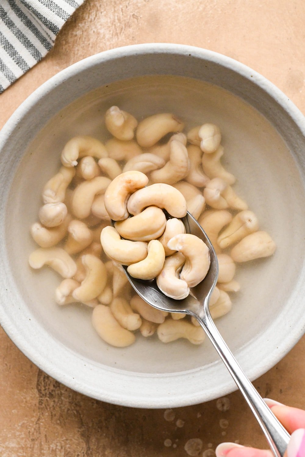 How to make dairy free hamburger helper: A spoon lifting cashews out of the bowl of soaking water to show what they look like after soaking for the right amount of time. Cashews are pale in color and plump.