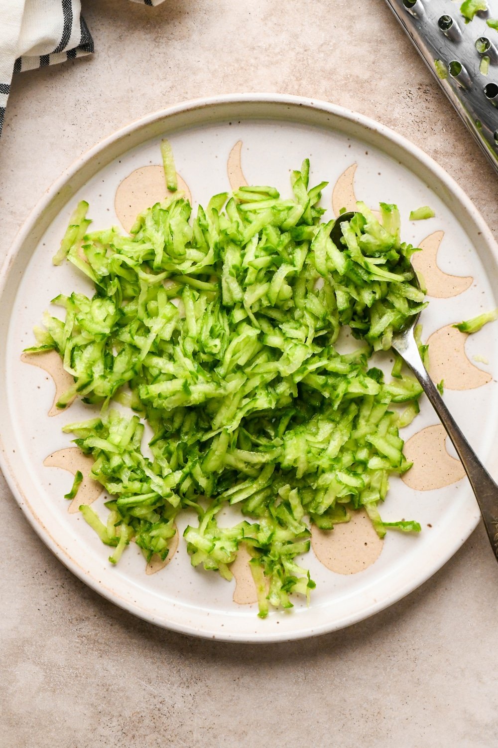 How to make dairy free tzatziki: Freshly grated cucumber on a beige colored ceramic plate with a spoon angled into the cucumber. A box grater that was used to shred the cucumber is next to the plate, along with a striped linen.