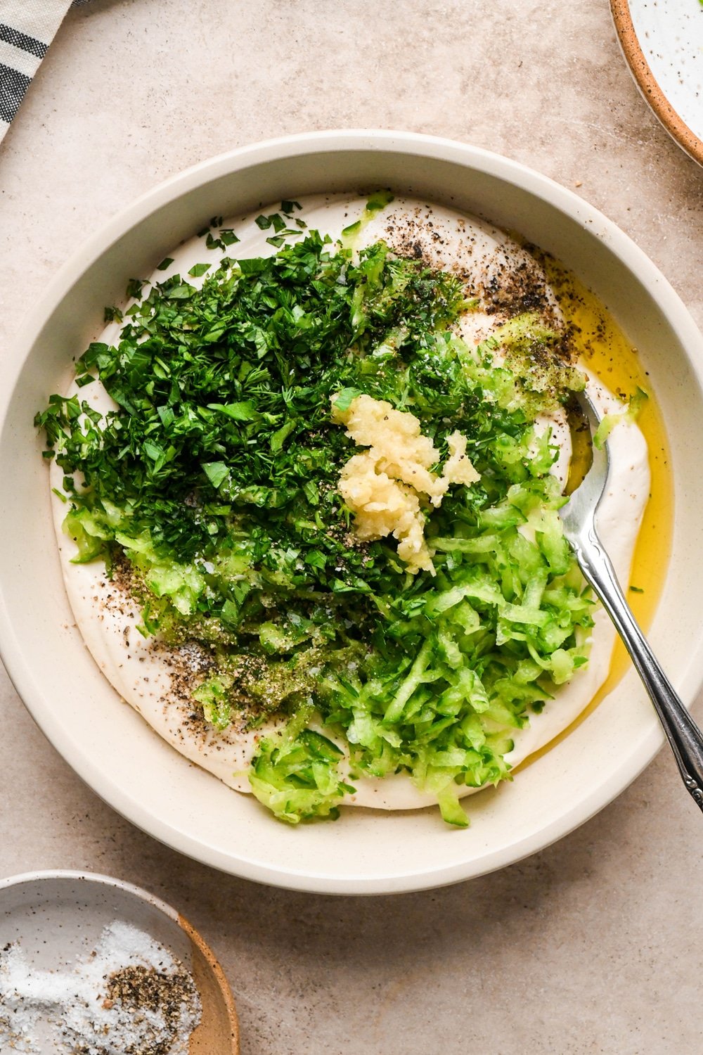 How to make dairy free tzatziki: Cashew cream, herbs, grated cucumber, garlic, salt, and pepper in a shallow cream colored bowl before stirring together to make tzatziki.