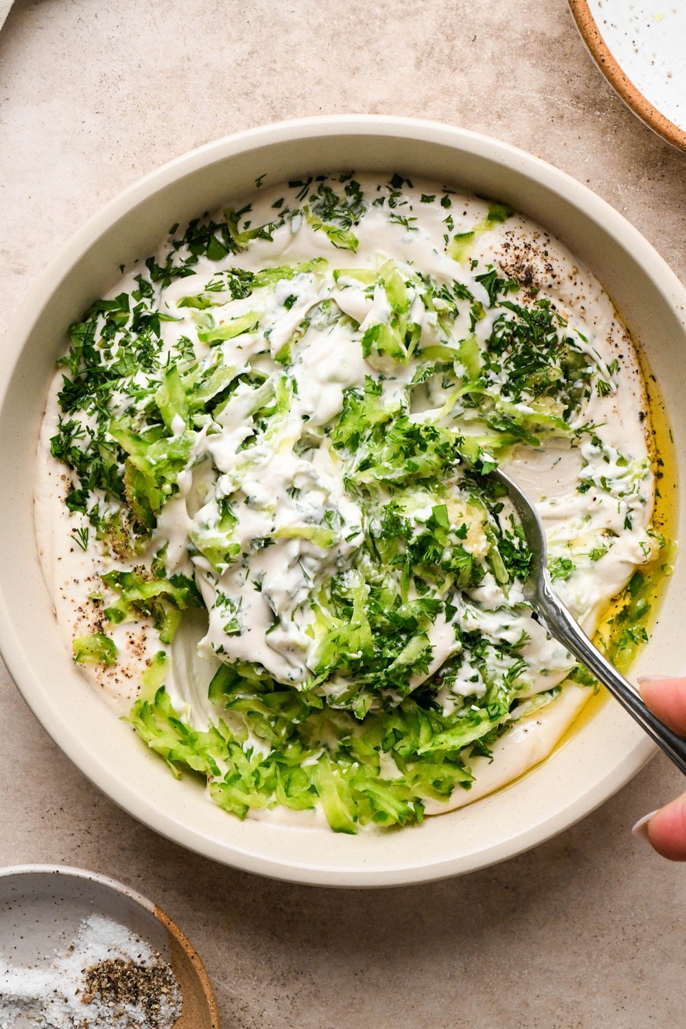 How to make dairy free tzatziki: Cashew cream, herbs, grated cucumber, garlic, salt, and pepper in a shallow cream colored bowl with a spoon stirring it all together to make tzatziki.