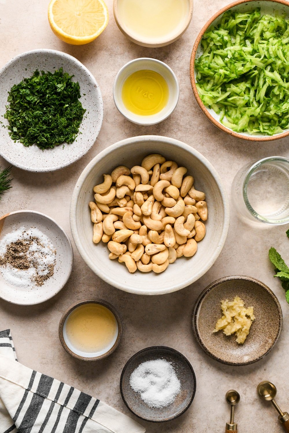 Ingredients for cashew cream tzatziki in various ceramics on a light cream colored background. There is a striped linen towel and wooden measuring spoons in the image as well.