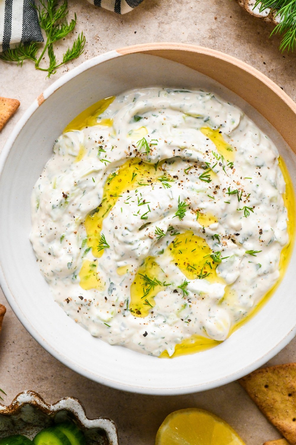 Creamy, dairy free tzatziki in a medium sized textured ceramic bowl. It is garnished with a drizzle of olive oil, black pepper, and chopped fresh dill.