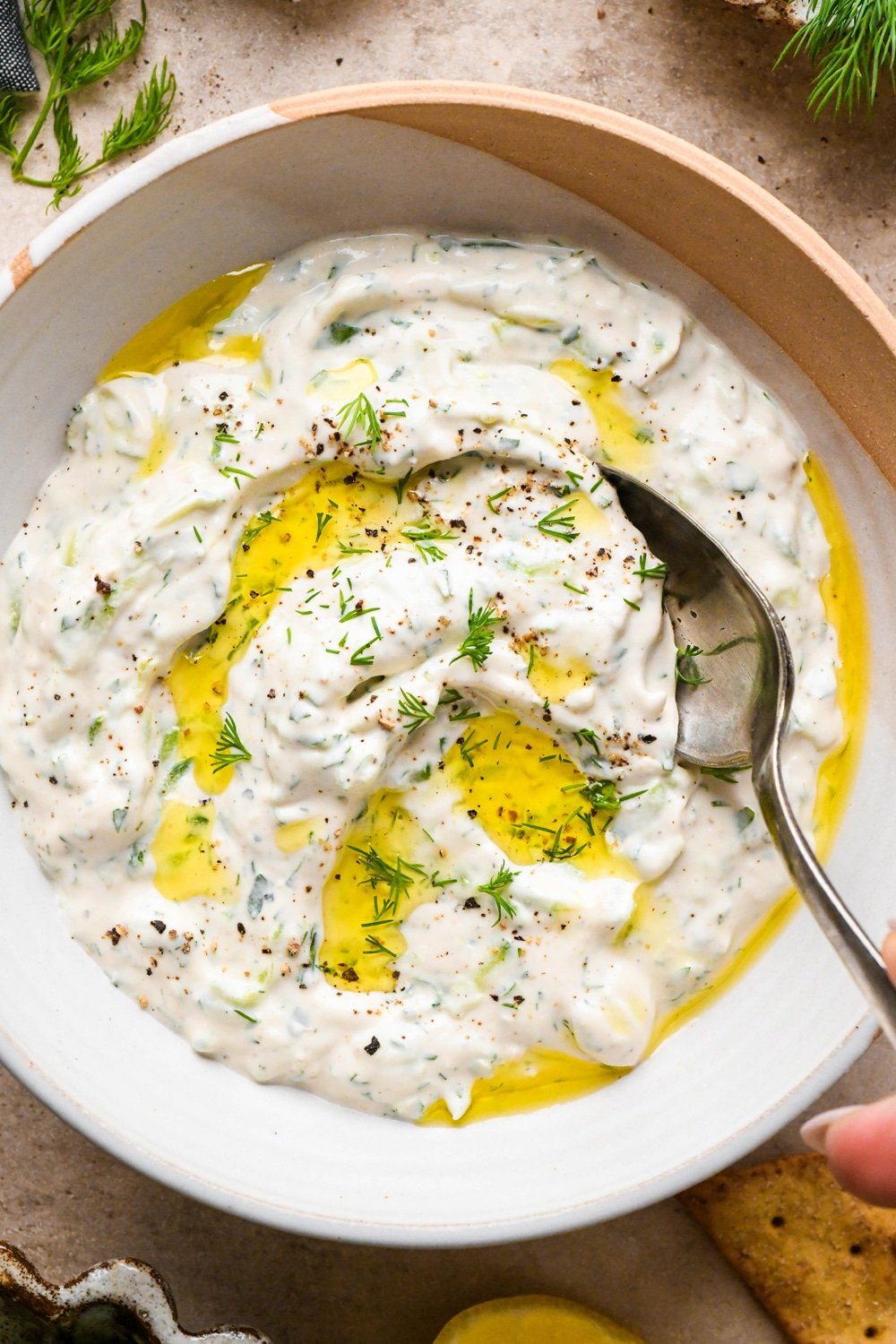 Creamy, dairy free tzatziki in a medium sized textured ceramic bowl. It is garnished with a drizzle of olive oil, black pepper, and chopped fresh dill. A spoon is dipping into the sauce to show the smooth texture.