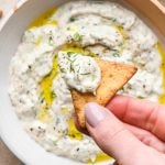 A hand holding a pita cracker that has been dipped into the tzatziki, over a bowl of the sauce.