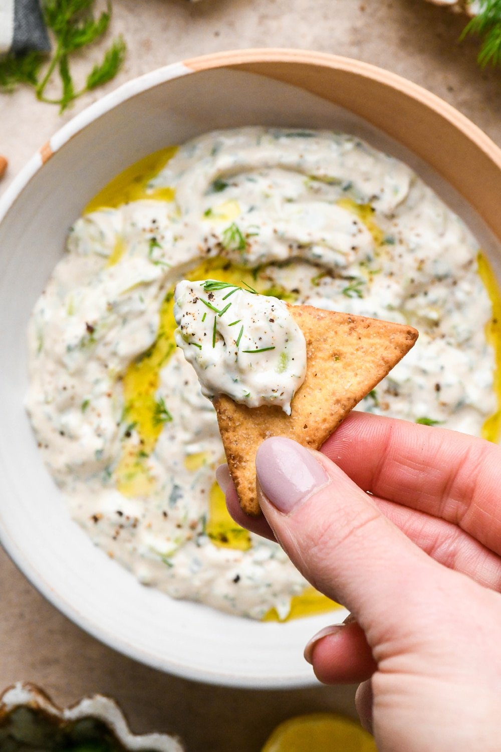 A hand holding a pita cracker that has been dipped into the tzatziki, over a bowl of the sauce. 