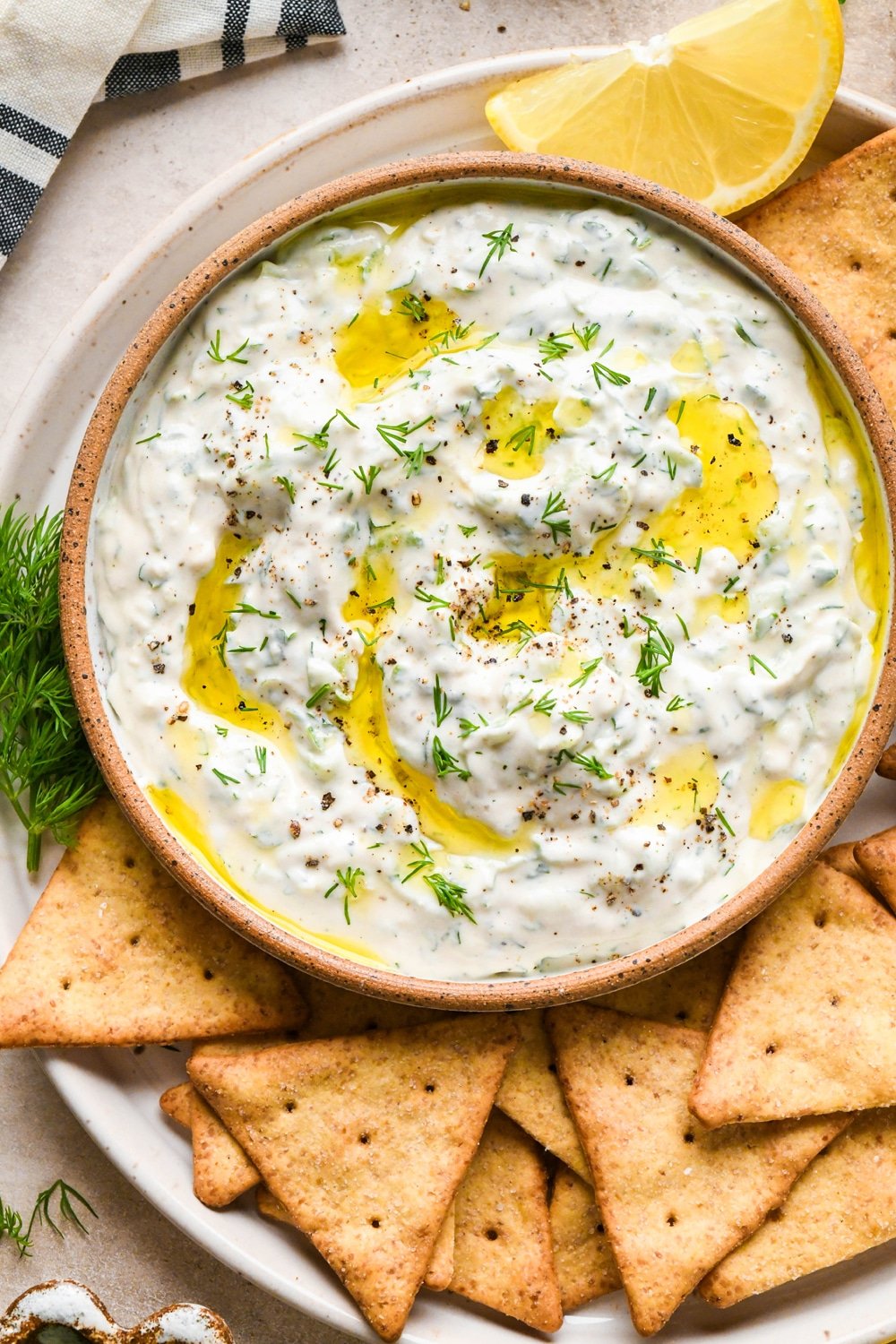 Dairy free cashew cream tzatziki in a small shallow ceramic serving dish, on a plate with gluten free pita crackers. Tzatziki is topped with a drizzle of olive oil, fresh dill, and cracked black pepper, and pieces of fresh dill surround the plate.