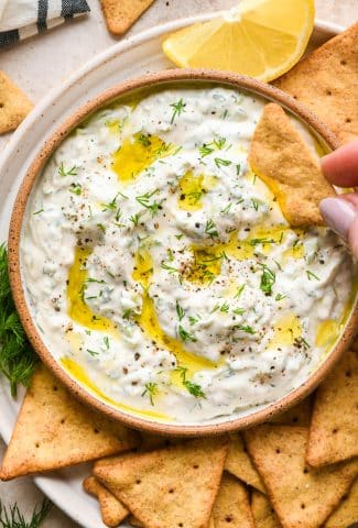 Dairy free cashew cream tzatziki in a small shallow ceramic serving dish, on a plate with gluten free pita crackers. Tzatziki is topped with a drizzle of olive oil, fresh dill, and cracked black pepper. A hand is dipping a cracker into the sauce.