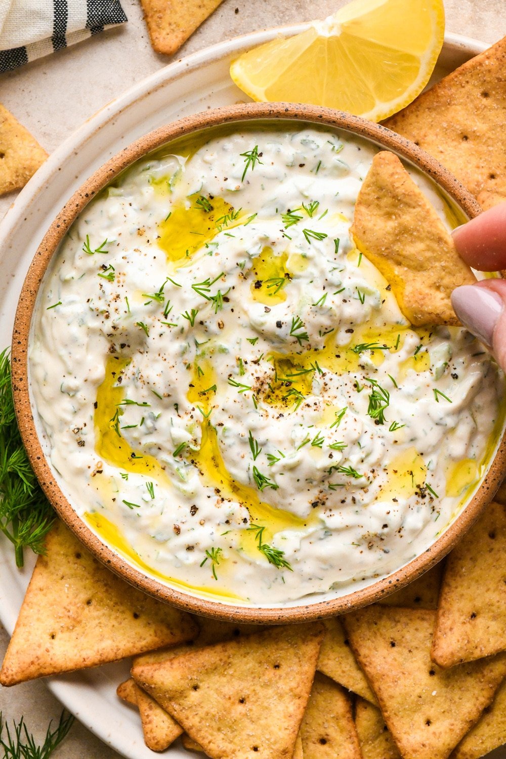 Dairy free cashew cream tzatziki in a small shallow ceramic serving dish, on a plate with gluten free pita crackers. Tzatziki is topped with a drizzle of olive oil, fresh dill, and cracked black pepper. A hand is dipping a cracker into the sauce.