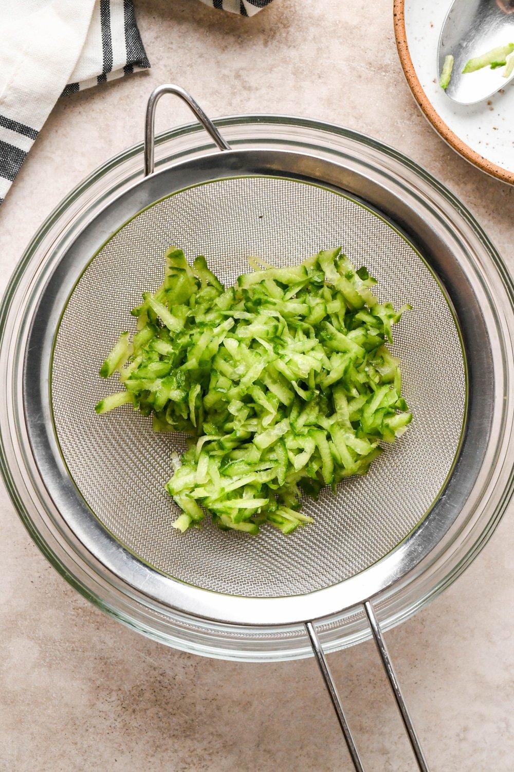 How to make dairy free tzatziki: Grated cucumber in a fine mesh strainer over a small clear glass bowl.