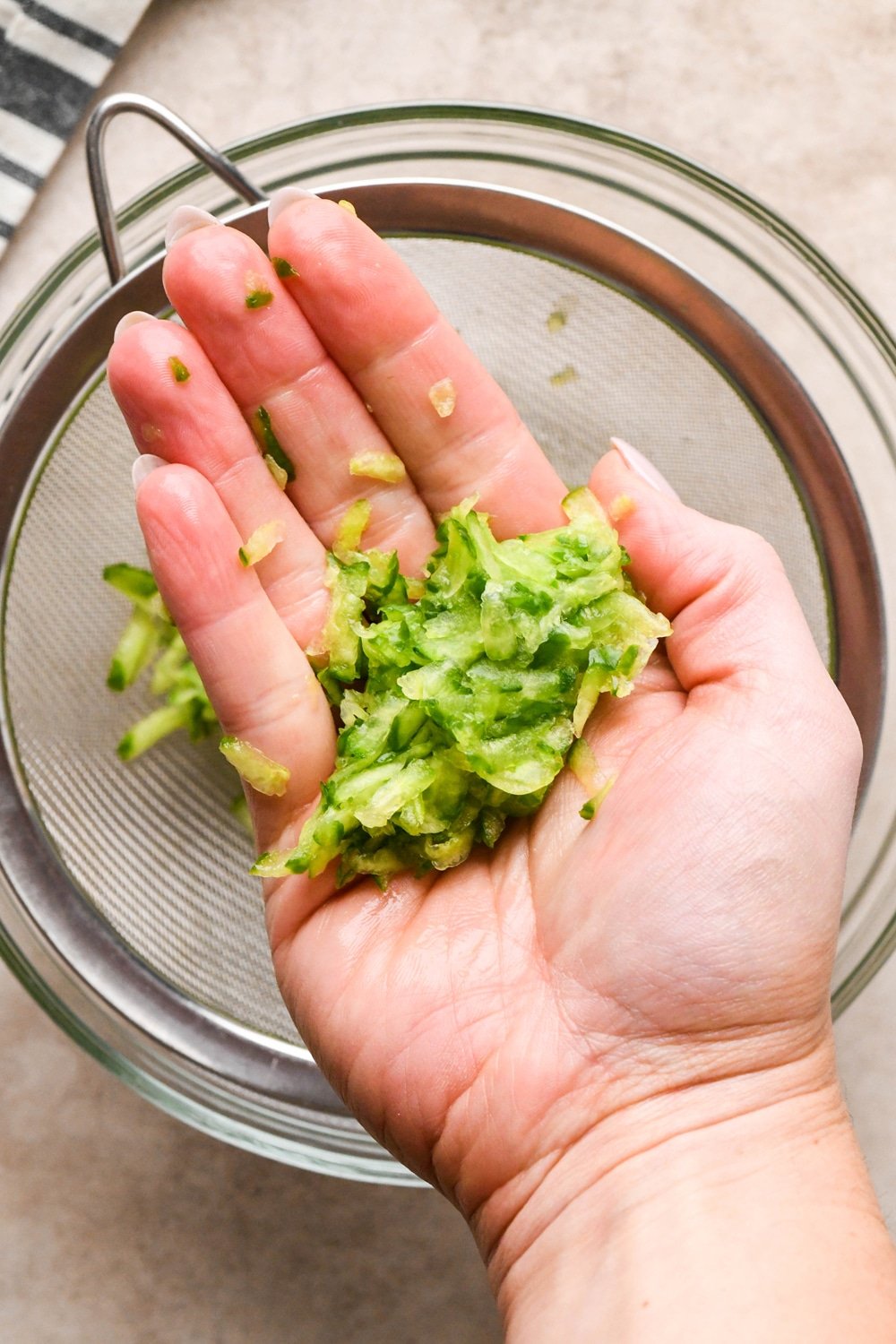 How to make dairy free tzatziki: A hand over the fine mesh strainer that contains grated cucumber holding a handful of cucumber that has been squeezed thoroughly to get rid of excess water.