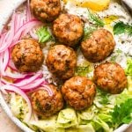 Baked Greek chicken meatballs in a shallow bowl with rice, cabbage salad, thinly sliced pickled red onions, creamy dairy free tzatziki sauce, and a lemon wedge. Bowl is garnished with scattered fresh dill and parsley, black pepper, and a tiny bit of aleppo chili flakes.