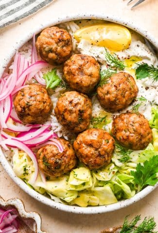 Baked Greek chicken meatballs in a shallow bowl with rice, cabbage salad, thinly sliced pickled red onions, creamy dairy free tzatziki sauce, and a lemon wedge. Bowl is garnished with scattered fresh dill and parsley, black pepper, and a tiny bit of aleppo chili flakes.